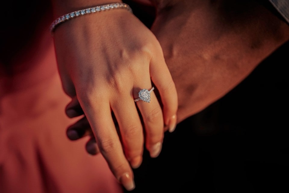 A woman donning a pear-shaped diamond halo ring and a diamond tennis bracelet holds her fiance’s hand.