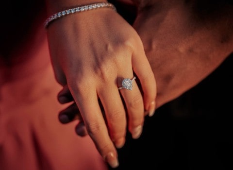 A woman donning a pear-shaped diamond halo ring and a diamond tennis bracelet holds her fiance’s hand.