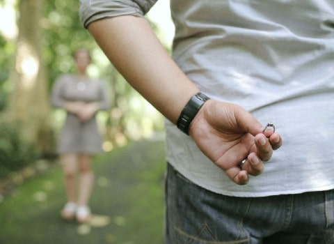 A man holds an engagement ring behind his back moments before proposing to his girlfriend during a picnic. 