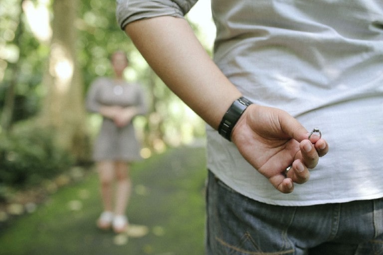 A man holds an engagement ring behind his back moments before proposing to his girlfriend during a picnic. 