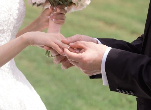 A groom slips a diamond wedding band on his bride’s ring finger during their outdoor wedding ceremony in the springtime.