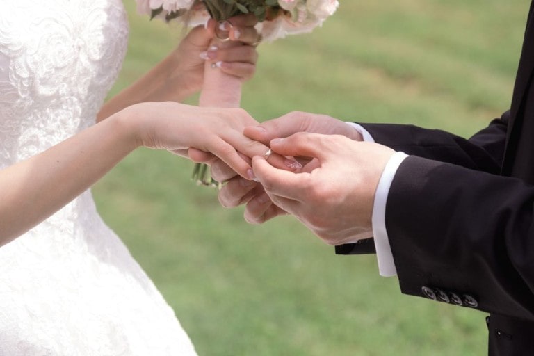 A groom slips a diamond wedding band on his bride’s ring finger during their outdoor wedding ceremony in the springtime.